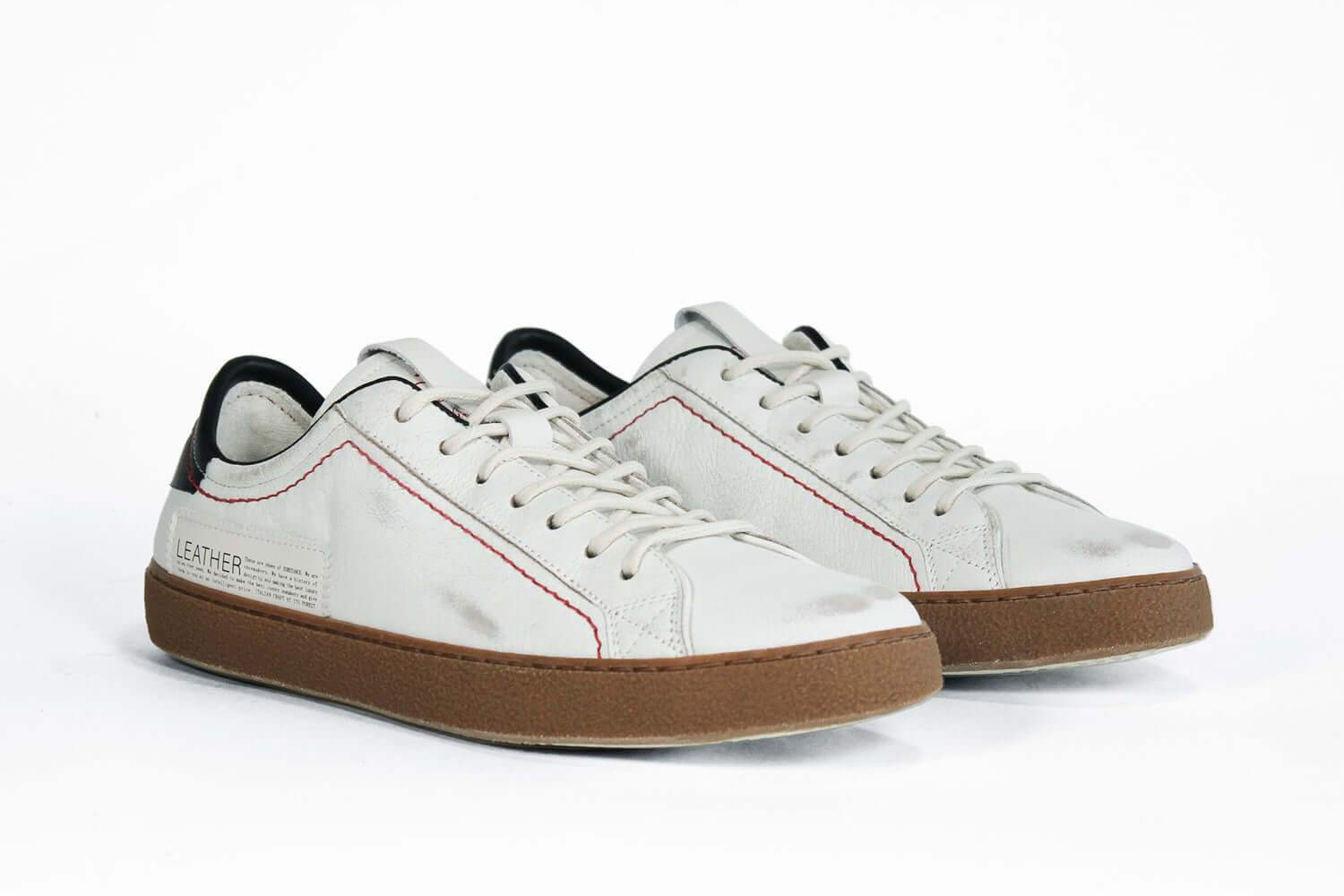 Three quarter front view of low top white sneaker with black accent. Distressed white leather upper with red stitching and dark rubber sole.