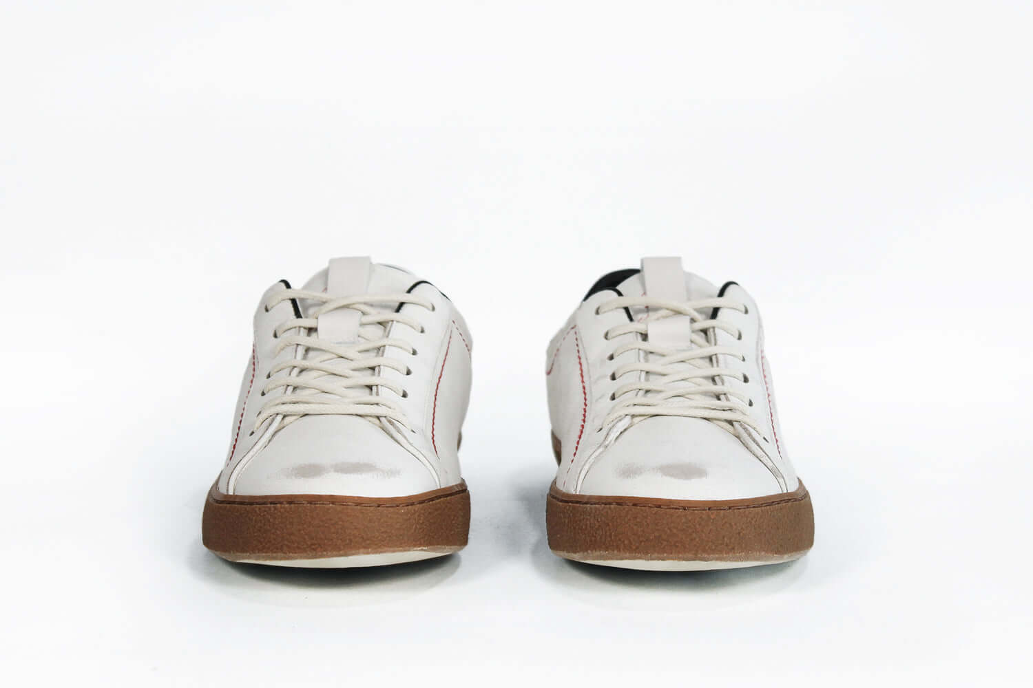 Front view of low top white sneaker with black accent. Distressed white leather upper with red stitching and dark rubber sole.