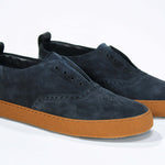 Three quarter front view of loafer style sneaker. Navy suede upper with perforated crown logo, elasticated tongue and a honey coloured recycled rubber sole.