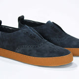 Three quarter front view of loafer style sneaker. Navy suede upper with perforated crown logo, elasticated tongue and a honey coloured recycled rubber sole.